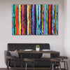 A large rectangular canvas wall art painting featuring a vibrant, multi-coloured strip abstract design of vertical brushstrokes, displayed on a light grey wall above a modern dining set with a dark grey sofa, wooden table, and stools.
