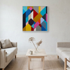 A square framed abstract geometric wall art painting with a colourful pattern of triangles and polygons, hanging on a light grey wall in a modern living room setting with a wooden coffee table and two beige sofas.