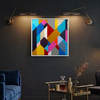 Geo Blocks 2 - A framed abstract geometric wall art painting with a white frame hangs on a dark charcoal wall in a styled living room setting, illuminated by two wall-mounted brass sconces. The artwork features a vibrant mix of blue, yellow, pink, orange, red, and gray shapes.