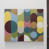 An abstract geometric canvas print featuring overlapping circles and semi-circles in muted colors including yellow, teal, olive green, pink, brown, blue, white, and maroon, displayed on a white wall.