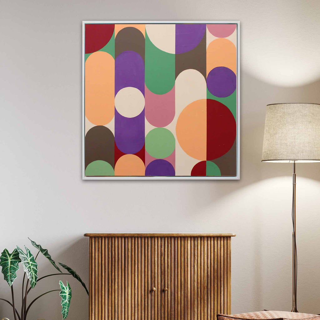 A square framed wall art painting with a mid-century modern abstract pattern of colourful vertical and circular shapes, hanging on a light beige wall above a wooden cabinet in a styled room with a floor lamp and plant.