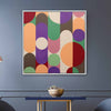 A square framed abstract art print featuring colorful geometric shapes like circles, ovals, and arcs in purple, green, orange, red, pink, brown, and cream, hanging on a slate blue-gray wall above a small table with a bowl.