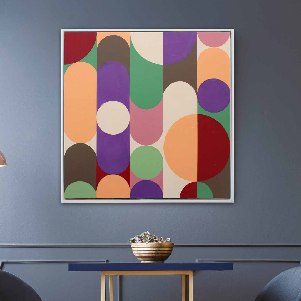 A square framed abstract art print featuring colorful geometric shapes like circles, ovals, and arcs in purple, green, orange, red, pink, brown, and cream, hanging on a slate blue-gray wall above a small table with a bowl.
