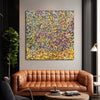 Rainbow Showers - A square abstract canvas painting featuring a dense, multi-coloured confetti or glitter pattern in yellow, blue, red, green, and orange, displayed on a light grey wall above a brown leather sofa in a modern living room setting.