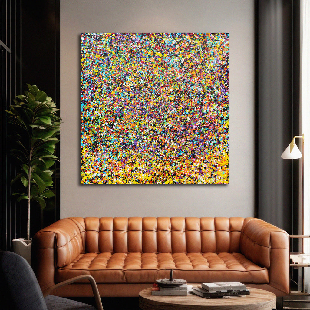 Rainbow Showers - A square abstract canvas painting featuring a dense, multi-coloured confetti or glitter pattern in yellow, blue, red, green, and orange, displayed on a light grey wall above a brown leather sofa in a modern living room setting.