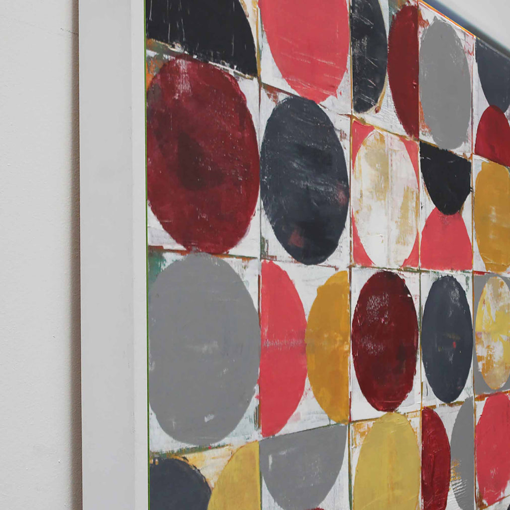 Side view of framed abstract geometric wall art featuring a grid of painted circles and half-circles in red, black, grey, Ocher, and white, shown hanging against a white studio wall.