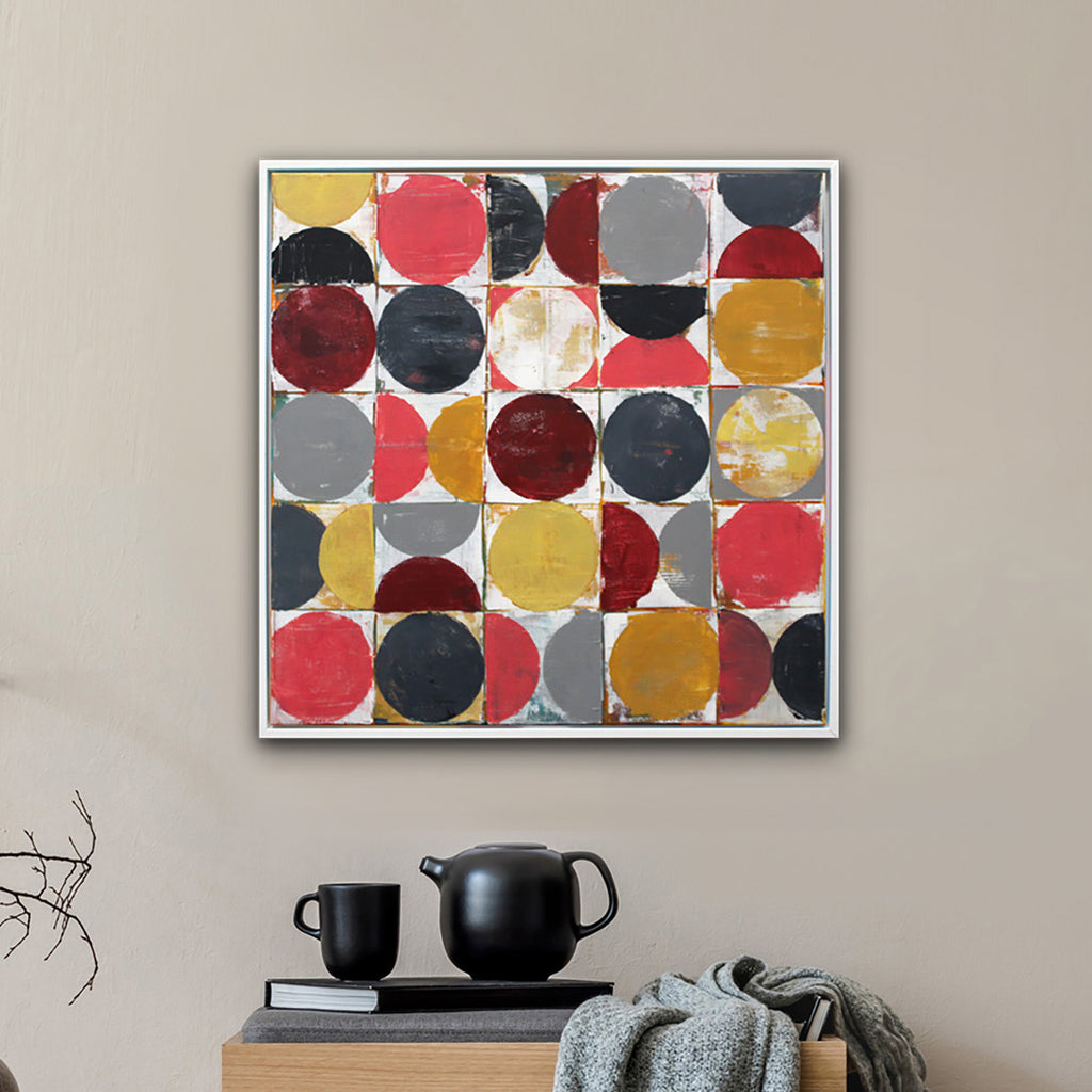 A framed abstract art painting featuring a grid of colourful circles in red, yellow, gray, and maroon, displayed on a beige wall above a wooden side table in a styled room setting.