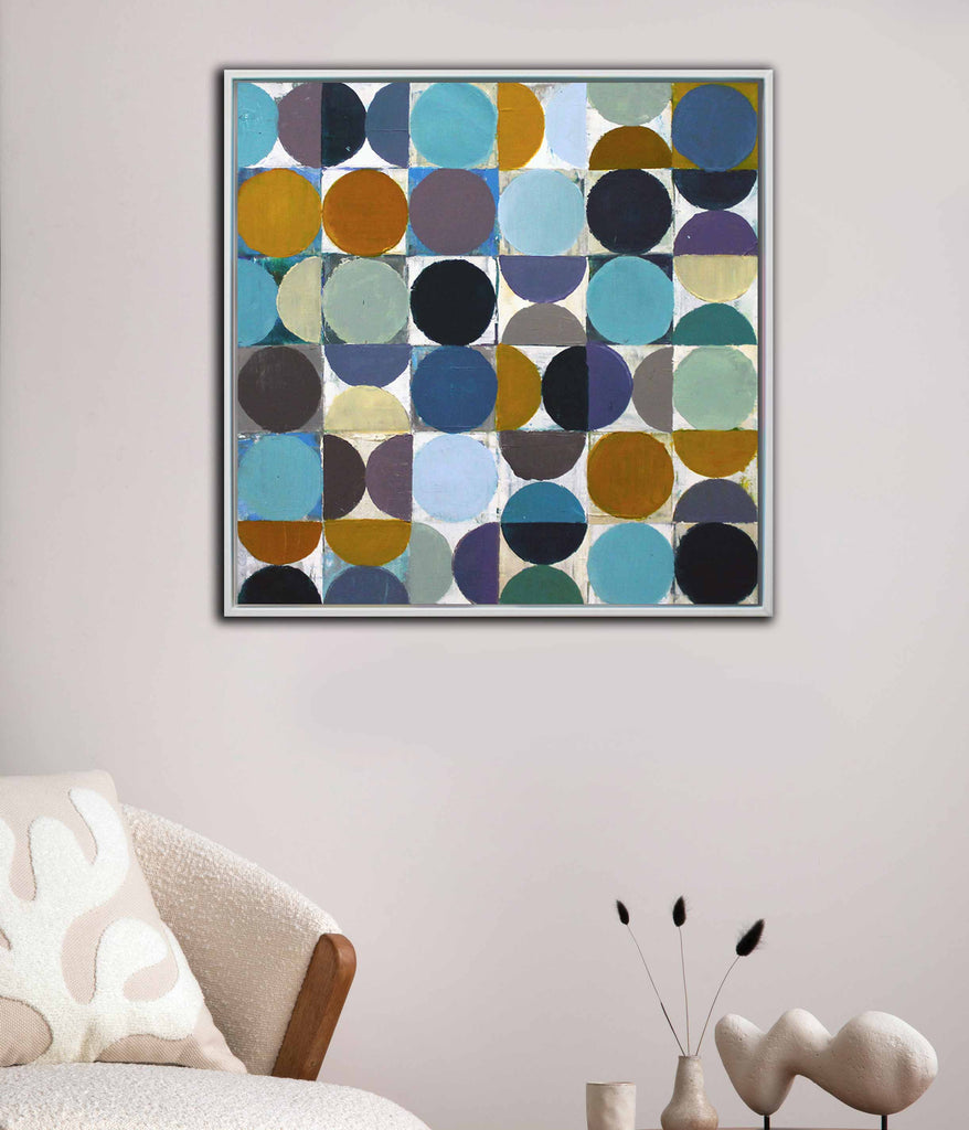 A white framed abstract geometric painting featuring a grid of circles and squares in shades of blue, teal, purple, brown, black, and mustard yellow on a light background hanging in a living room setting.