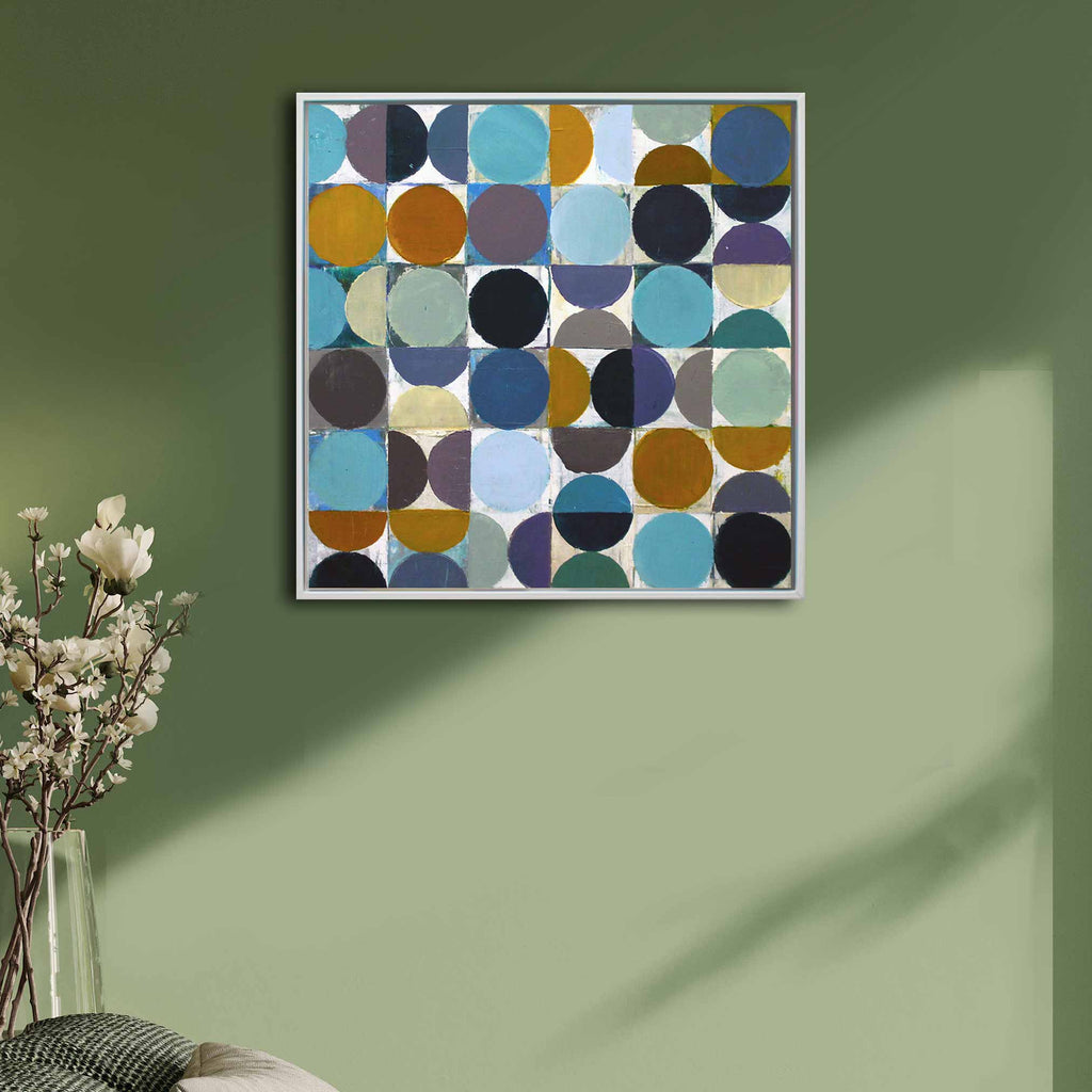 A white framed abstract spot painting featuring white squares and contrasting circles in shades of blue, teal, purple, brown, black, and mustard hanging on a living room wall.