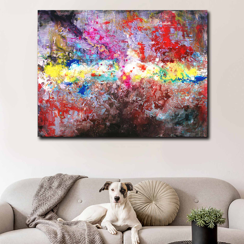 A large rectangular abstract canvas painting with bold splashes of red, pink, blue, yellow, white, and dark tones hangs on a light beige wall above a sofa with a dog lying on it and a small potted plant in the foreground.