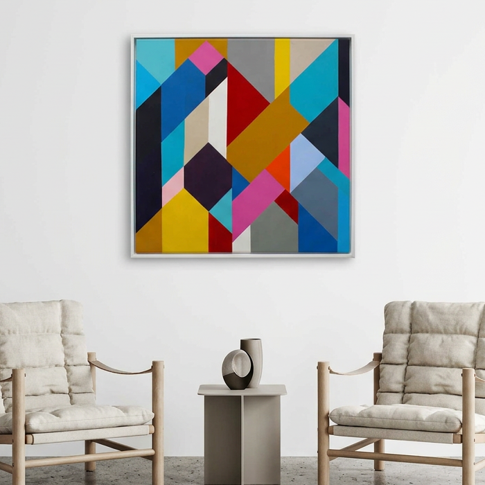 Geo Blocks No 2 - A square framed abstract geometric wall art painting with colourful overlapping shapes in blue, yellow, red, pink, grey, and black, displayed above two light beige armchairs in a minimalist room setting.