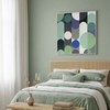 A styled bedroom scene with a bed dressed in sage green and white linens beneath a framed abstract geometric wall art painting. The room has a pale green wall, a woven rattan headboard, white curtains, and a small table with a vase and books.