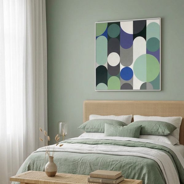 A styled bedroom scene with a bed dressed in sage green and white linens beneath a framed abstract geometric wall art painting. The room has a pale green wall, a woven rattan headboard, white curtains, and a small table with a vase and books.