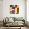 A framed abstract wall art painting with overlapping vertical oval and circular shapes in mustard yellow, teal blue, coral pink, rust red, tan, and cream, hanging above a green sofa in a bright living room.