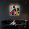 Geo Blocks 1 - A framed abstract geometric wall art painting with colourful polygon shapes hangs above a dark velvet sofa in a dimly lit room. A white and black dog lies on the sofa, and a small round side table with a potted plant is in the foreground.