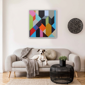 Geo Blocks 1 - A colourful geometric abstract wall art print hangs above a light grey sofa in a styled living room. A white dog lies on the sofa, and a black woven side table with a small plant sits in front.