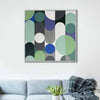 A white framed abstract geometric wall art painting with overlapping rounded shapes in green, blue, purple, grey, white, and black, displayed above a light blue sofa in a living room setting.