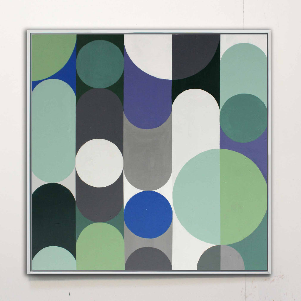 abstract art painting hanging in a studio wall featuring large green, blue, grey, and white geometric circles on a white background framed in a white floating frame.