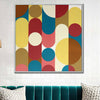 A square framed abstract wall art painting with bold geometric shapes in yellow, blue, red, pink, cream, and brown, displayed above a teal velvet sofa against a white brick wall.