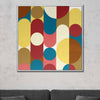 A square framed abstract wall art painting with bold geometric shapes in yellow, red, blue, cream, and brown, displayed on a light gray textured wall above a dark gray sofa.