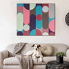 A square canvas wall art print with a colorful geometric pattern of vertical bars and circles in pink, teal, gray, brown, and cream, displayed on a white wall above a beige sofa with a dog lying on it.