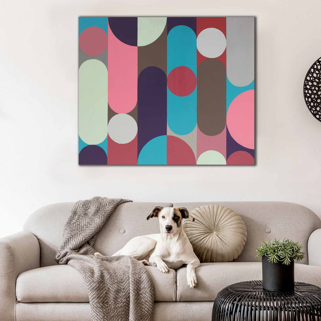 A square canvas wall art print with a colorful geometric pattern of vertical bars and circles in pink, teal, gray, brown, and cream, displayed on a white wall above a beige sofa with a dog lying on it.