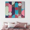 A square abstract wall art painting with vertical rounded shapes in pink, teal, brown, cream, and grey, displayed above a bed with dusty rose and white pillows in a bedroom setting.
