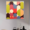 A square framed wall art painting featuring a mid-century modern abstract pattern of overlapping vertical arches and circles in olive green, red, blue, cream, and orange, hanging on a light gray wall above a table with decorative objects.