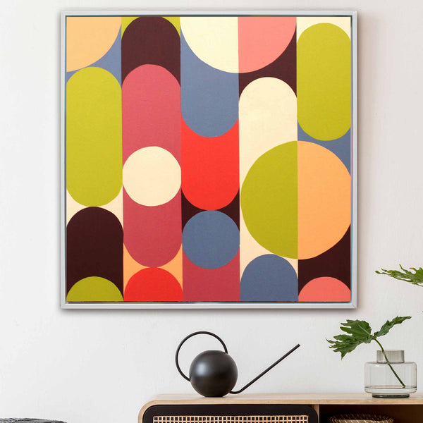 A square framed abstract wall art print with bold mid-century geometric shapes in red, yellow, blue, pink, cream, and brown, hanging on a white wall above a console table with a black watering can and a small plant.