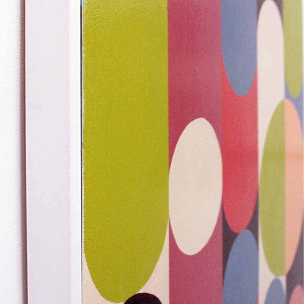 Close-up of a framed abstract art print featuring bold vertical bands of olive green, pink, cream, blue, and peach circles on a white frame.