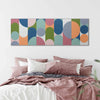 A colorful abstract wall art painting with overlapping arches and circles in muted blue, green, pink, orange, and gray tones, displayed above a bed with dusty rose bedding in a softly lit bedroom.