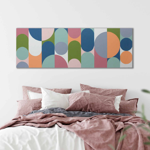 A colorful abstract wall art painting with overlapping arches and circles in muted blue, green, pink, orange, and gray tones, displayed above a bed with dusty rose bedding in a softly lit bedroom.