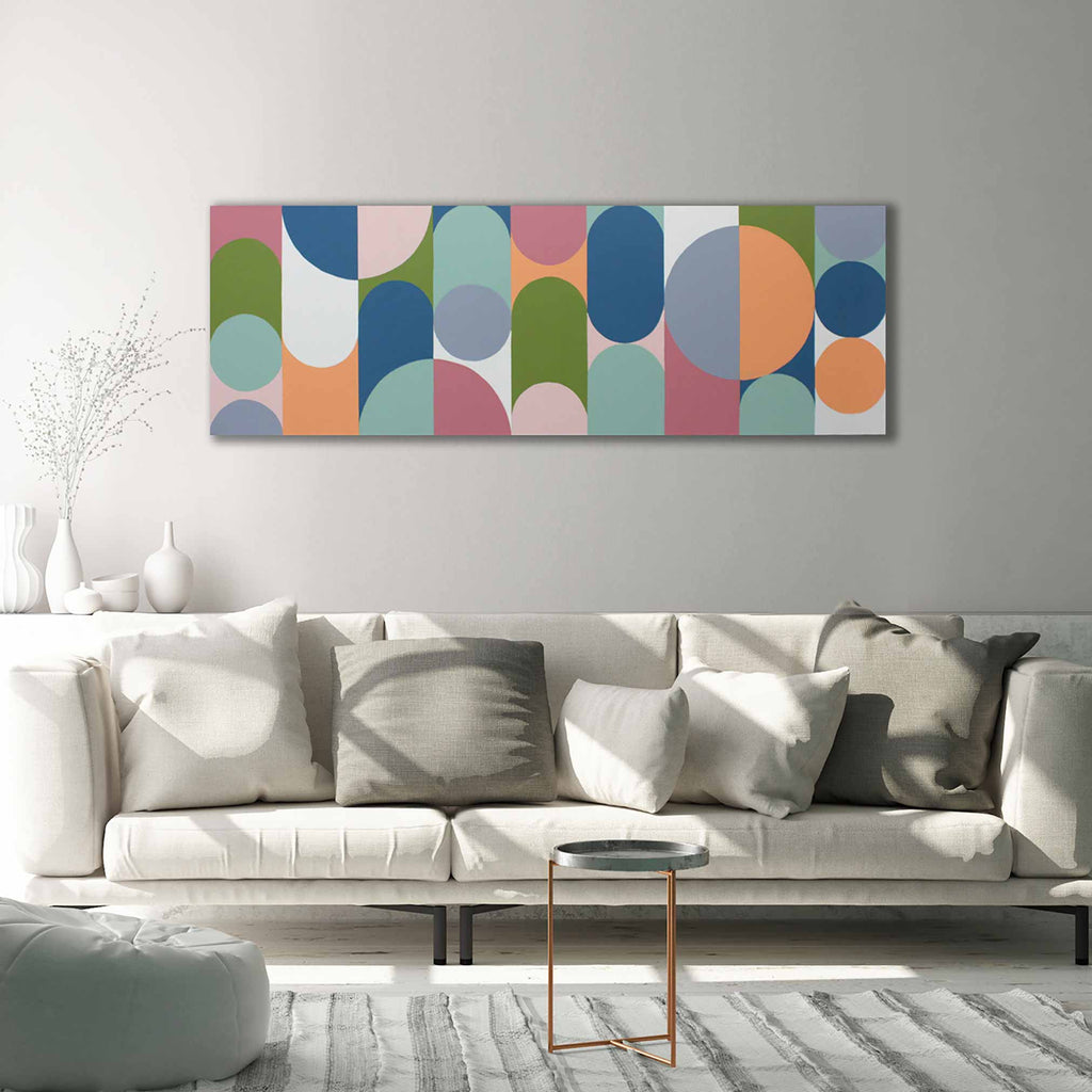 A wide rectangular abstract wall art piece with overlapping semicircles and arcs in muted blue, teal, pink, orange, green, and cream, displayed above a light beige sofa in a bright modern living room.
