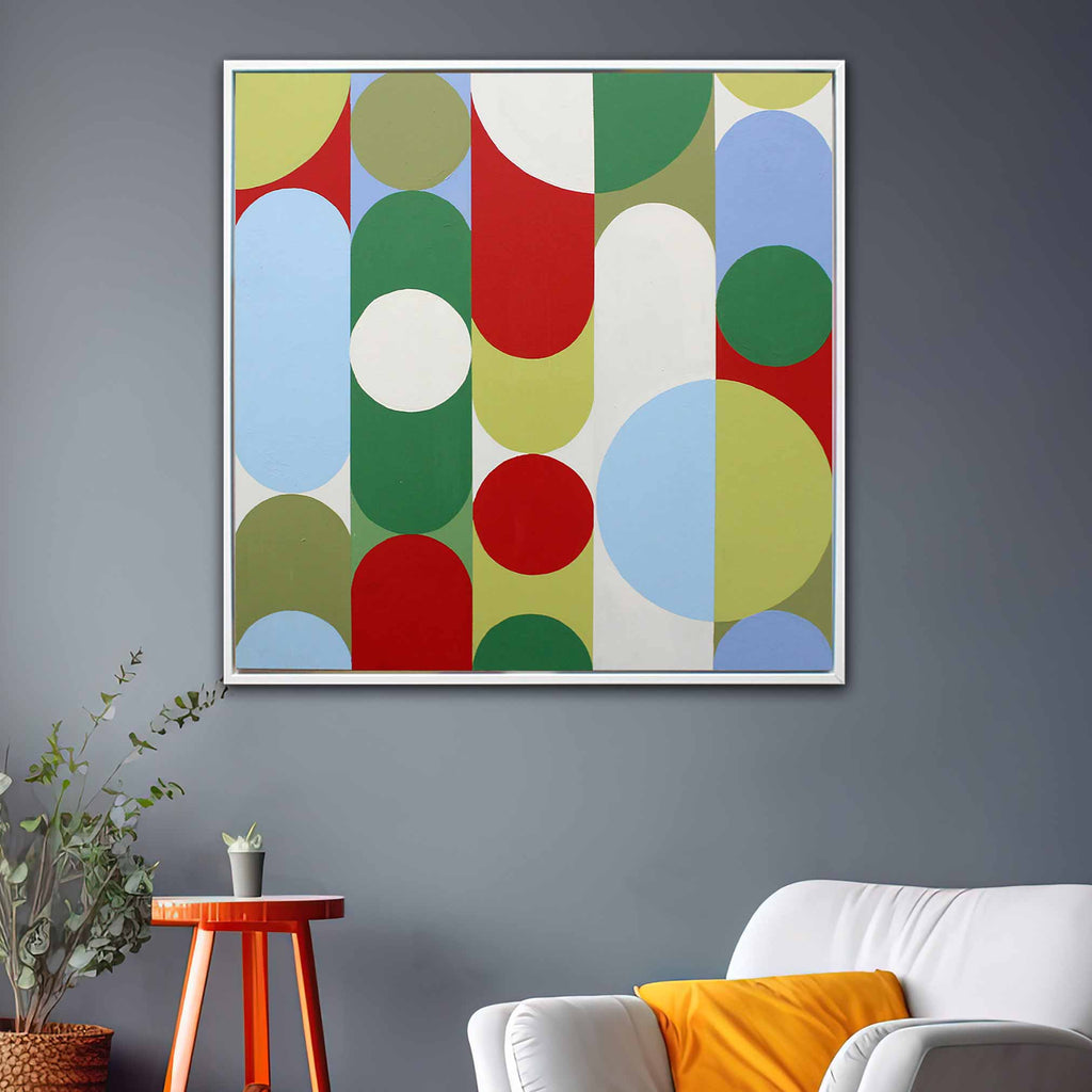 A square white framed abstract wall art painting with overlapping circles and arcs in red, green, blue, yellow, white, and olive tones, displayed on a dark gray wall above a white sofa with an orange pillow, next to a small wooden side table with a plant.