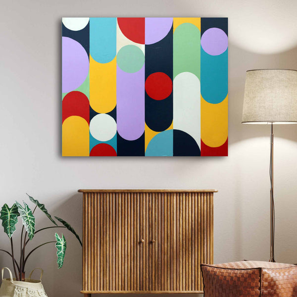 A square canvas wall art painting featuring a colourful mid-century modern geometric pattern of overlapping circles and arches, displayed on a light grey wall above a wooden cabinet in a styled room setting.