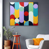 A square abstract wall art print with vertical bars and overlapping circles in yellow, teal, purple, red, black, white, and mint, displayed on a dark gray wall above a white armchair with an orange pillow, next to a small orange stool and potted plants.