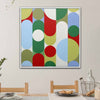 A square framed abstract wall art painting hangs on a light beige wall above a dining table. The artwork features large overlapping circles and arcs in green, red, light blue, olive, and white.