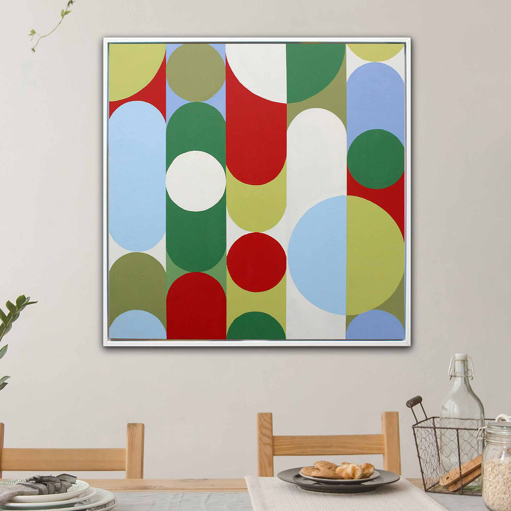 A square framed abstract wall art painting hangs on a light beige wall above a dining table. The artwork features large overlapping circles and arcs in green, red, light blue, olive, and white.