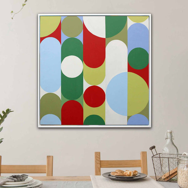 A square framed abstract wall art painting hangs on a light beige wall above a dining table. The artwork features large overlapping circles and arcs in green, red, light blue, olive, and white.