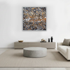 A square abstract wall art painting with a white frame hangs above a modern light grey sideboard in a minimalist living room. The artwork features a dense, textured pattern of black, grey, white, and warm orange-brown splatters.
