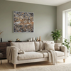 A square framed abstract wall art painting with a textured, metallic-looking pattern in gold, bronze, grey, and white, hanging on a sage green wall above a beige sofa in a modern, well-lit living room.
