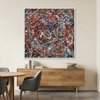 A framed abstract painting with a dense splatter pattern of red, blue, black, white, and yellow paint hangs on a beige wall in a modern dining room setting with a wooden sideboard, dining table, and gray chairs.