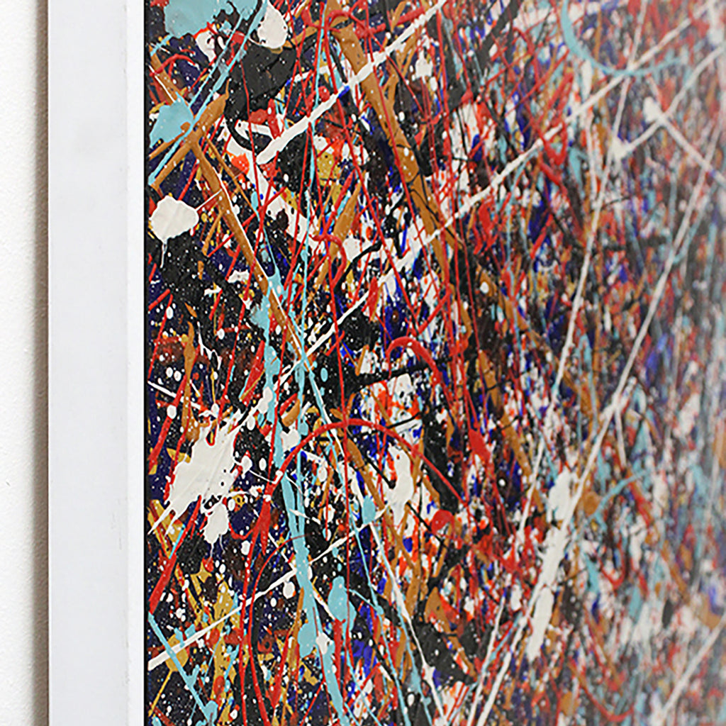 Side view close-up of a framed abstract drip-style painting with energetic splatters and drips in red, blue, white, black, and gold tones, shown against a white wall.