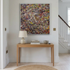A large framed abstract painting with dense multicolored brushstrokes hangs on a light gray wall above a wooden console table in a bright, modern interior.