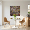 A large framed abstract canvas painting with a dense multi colour paint splatter pattern hangs on a white wall in a bright dining room. The room features a white tulip table, two tan Scandinavian design chairs, and light stone tile flooring.