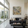 A large abstract canvas painting with splatters of orange, yellow, black, white, and green, displayed on a beige wall above a modern gray sofa in a bright, contemporary living room.