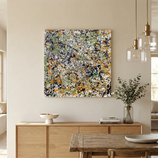 A large square abstract canvas painting with a dense paint-splatter pattern in white, orange, green, black, and grey, displayed on a beige wall above a light wood console table in a modern dining room setting.