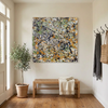 A large square abstract canvas painting with a dense splatter pattern in black, white, orange, green, and grey hangs on a beige wall in a bright entryway. A light wood bench with a cream throw sits below, and a potted olive tree stands to the left.