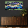 An abstract landscape canvas painting called Malva Wave inspired by The North East Coastline with vibrant blues, purples, greens, and gold brushstrokes, displayed on a dark grey wall above a mid-century modern wooden sideboard with a potted fern in a gold vase.