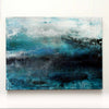 An abstract canvas painting featuring textured brushstrokes of teal, blue, black, and white, with a hazy, atmospheric composition displayed on a plain white wall in the artists studio.